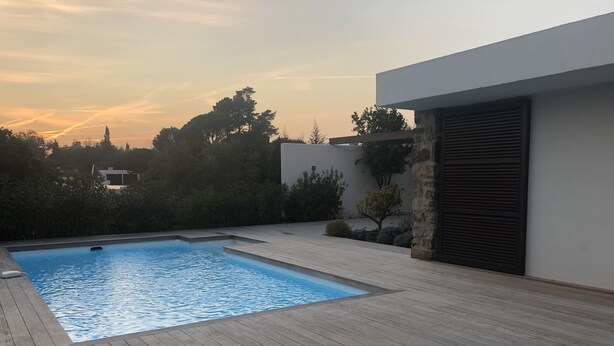 Une piscine rectangulaire entourée d’une terrasse en bois, avec en arrière-plan des arbres et un bâtiment moderne.