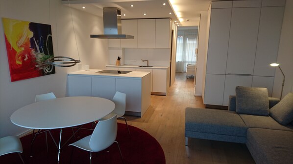A modern living room with a grey sofa, a round dining table with white chairs, a kitchen area with white cabinets, and a red rug.