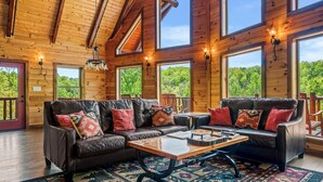 A wooden cabin interior with a leather sofa, a coffee table, and large windows offering a forest view. - Ridge View Lodge • Nature/Mtn View • Hot tub • Firepit • Games • Parking 6 cars (Sevierville)