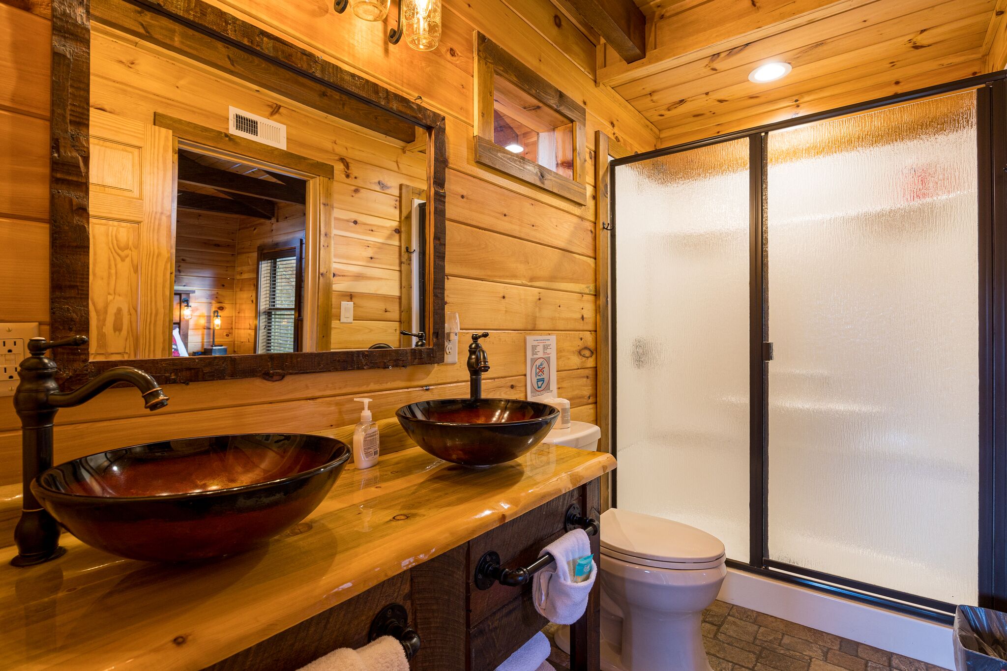 A wooden bathroom with a large mirror, two sinks, and a toilet.