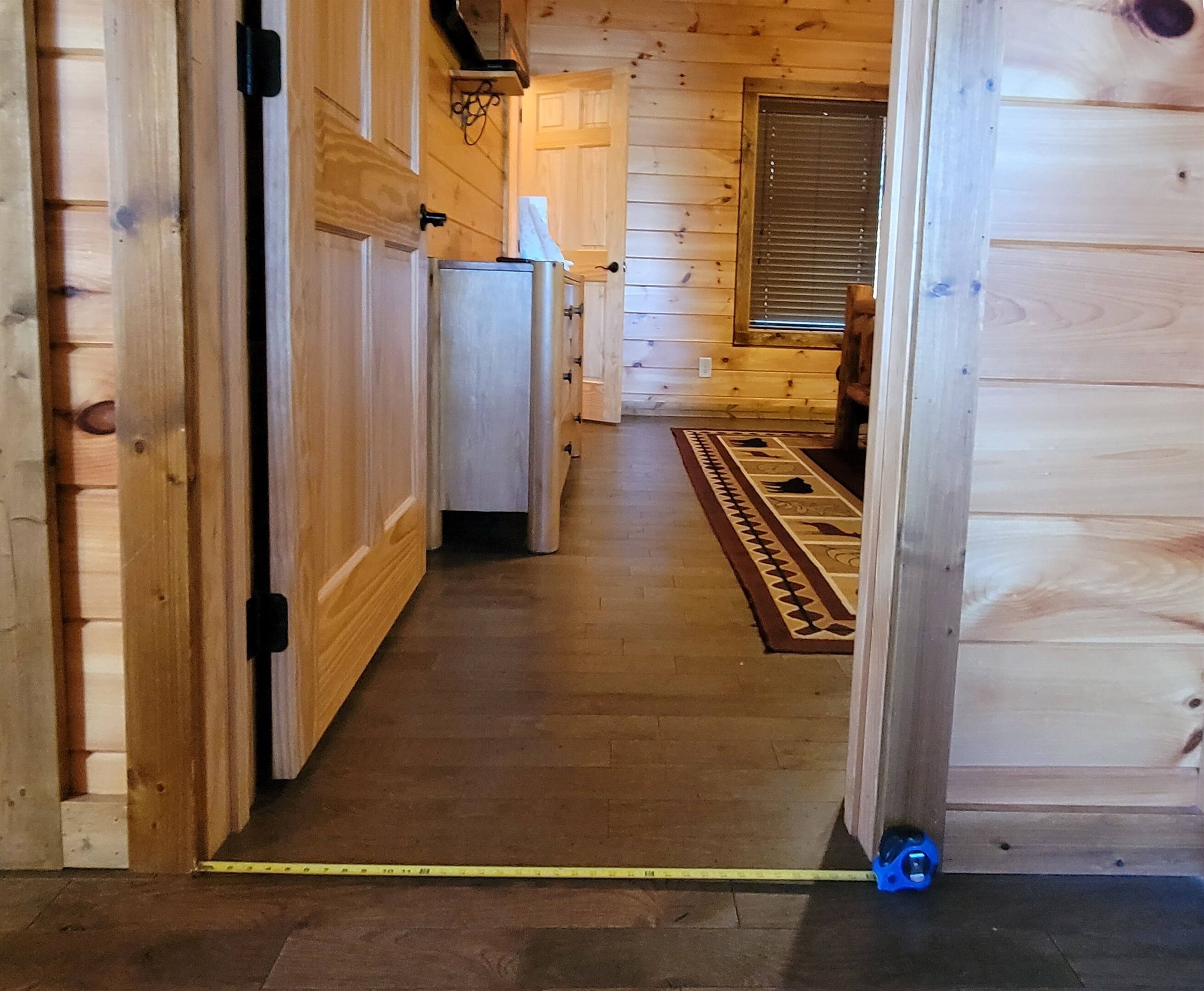 Een houten hal met een deur naar een kamer, een raam met jaloezieën en een kamerbreed tapijt op de vloer.