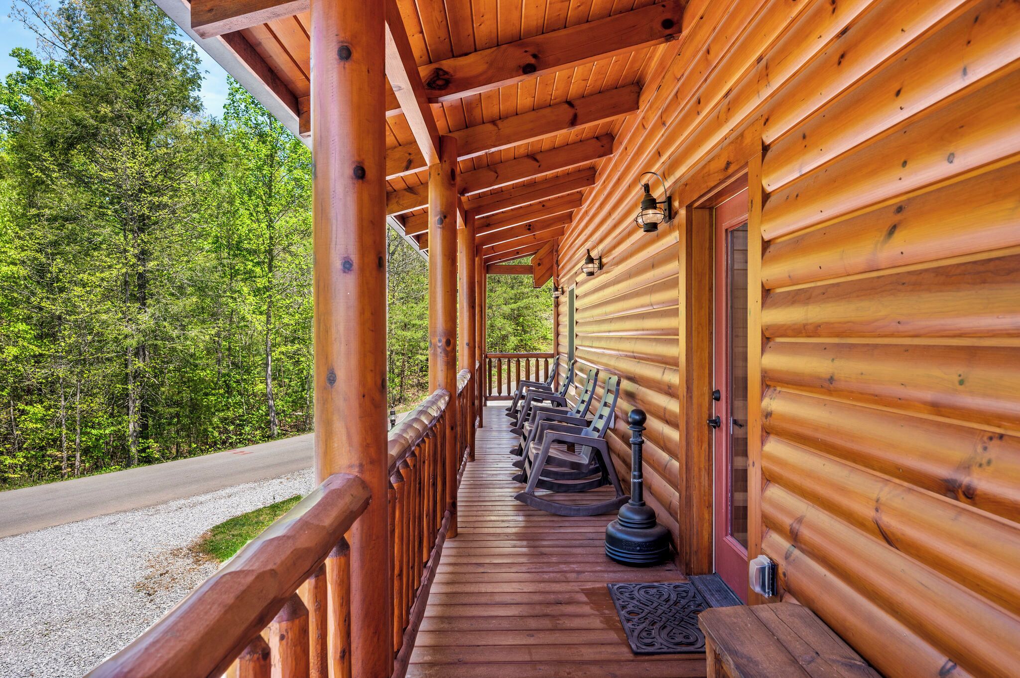 A wooden cabin with a covered porch, wooden benches, and a view of a forested road.