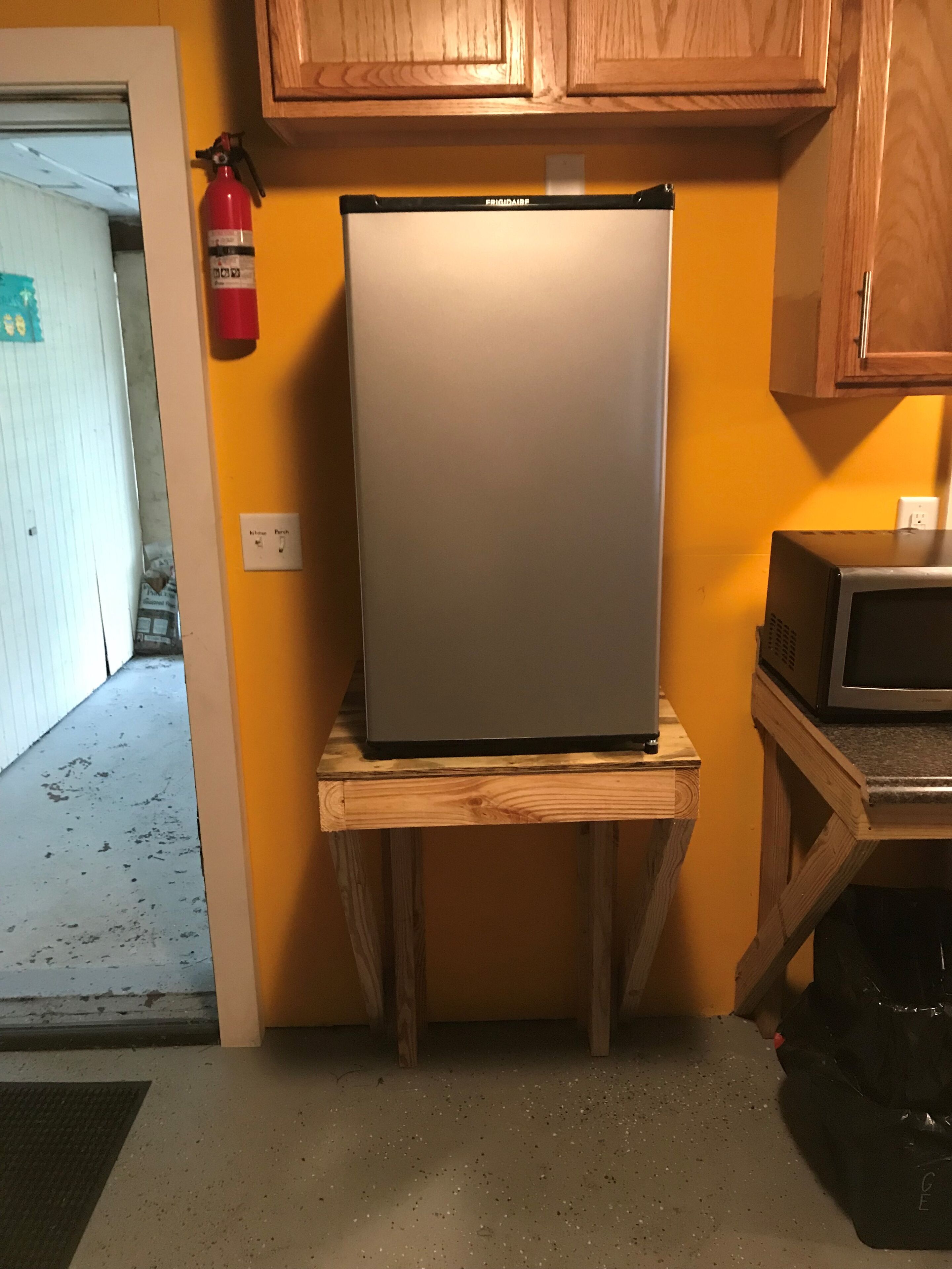 Una cocina con un refrigerador sobre un soporte de madera, un microondas y un extintor en la pared.