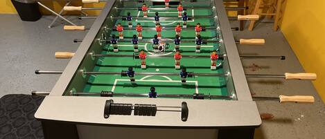 Une table de baby-foot dans une pièce avec un mur jaune, une fenêtre et une poubelle.