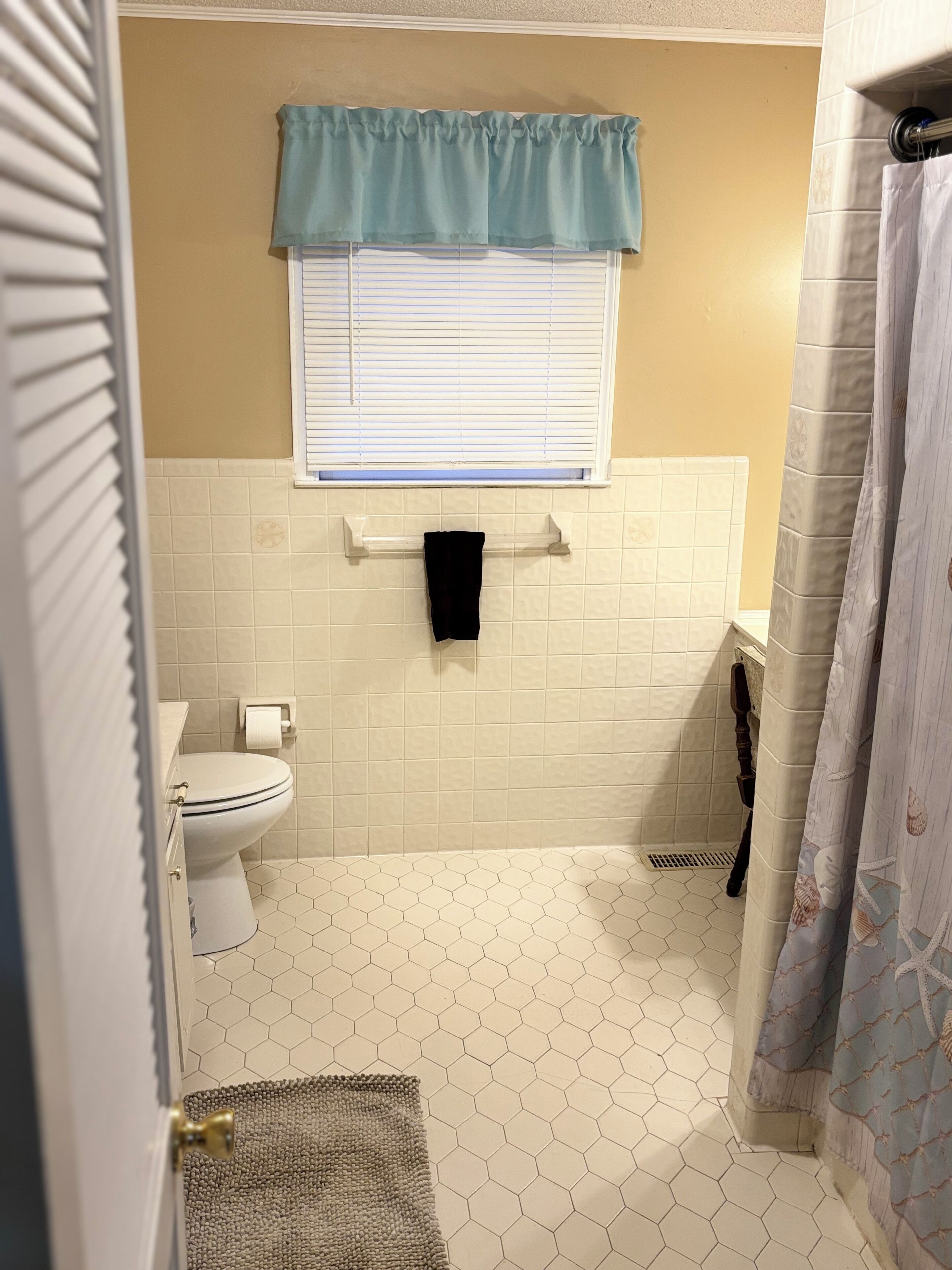 A bathroom with a toilet, a towel rack, a window with blinds, and a shower curtain.