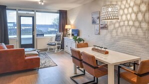 Ein modernes Wohnzimmer mit Sofa, einem Esstisch mit Stühlen, einem Fernseher und einer Lampe. Ein großes Fenster mit Vorhängen bietet Blick auf die Gebäude draußen.