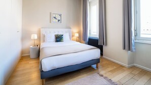Un dormitorio con cama, lámparas de noche, una ventana con cortinas, un radiador y un cuadro colgado en la pared.