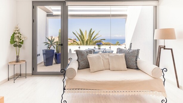 Een slaapkamer met een bed, een tafel met stoelen, een lamp en uitzicht op zee door een schuifdeur.