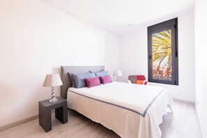 A bedroom with a bed, two bedside tables, a window with a view of palm trees, and a door.