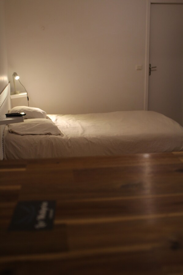 Un lit simple avec un matelas et des oreillers blancs, une table de chevet avec une lampe et un plancher en bois.