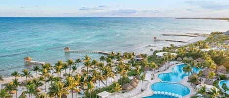 A resort with a large swimming pool, palm trees, and a pier extending into the ocean.