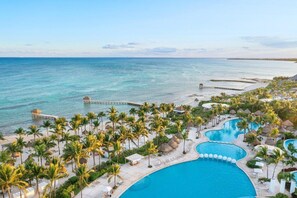Ein Resort mit einem großen Schwimmbad, Palmen und einem Steg, der ins Meer hinausführt.