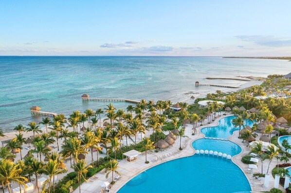 A resort with a large swimming pool, palm trees, and a pier extending into the ocean.
