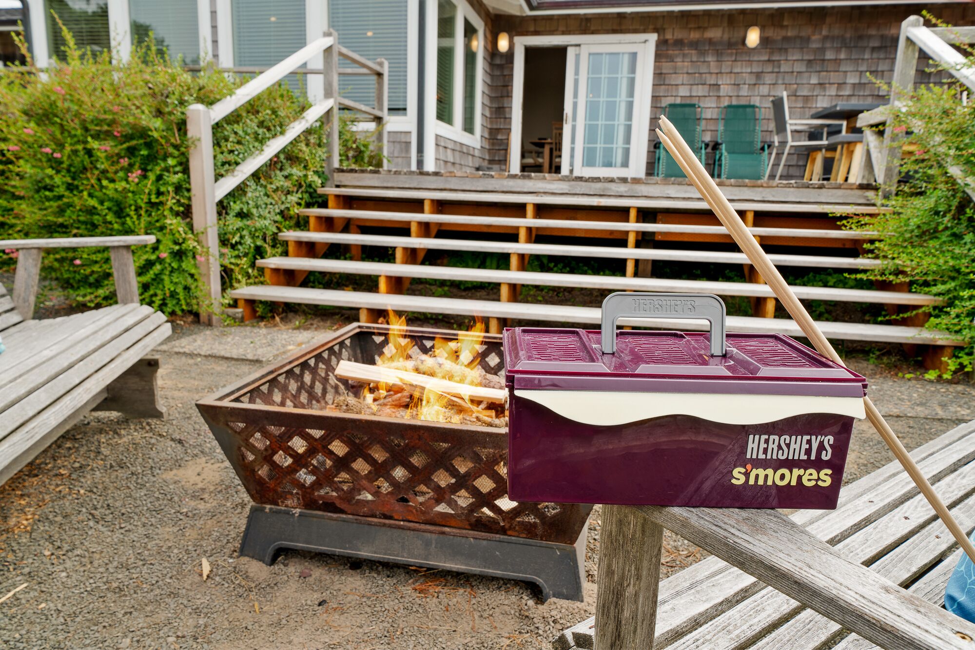 Een draagbare grill met een doos Hershey's S'mores er aan bevestigd, buiten geplaatst met houten banken en een huis op de achtergrond.