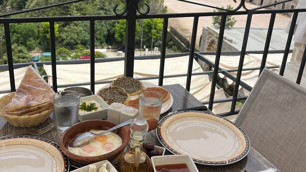 A balcony with a table set for breakfast, overlooking a historic building and lush hills. - Beit Lebbos (Beit Al Dine)