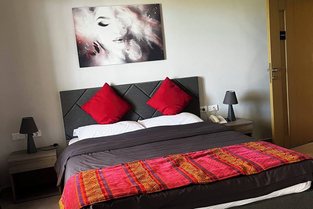 Un dormitorio con una cama, una mesita de noche con lámpara, un cuadro en la pared y un equipo de aire acondicionado.