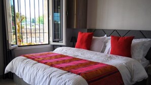 A bed with a white comforter and a colorful blanket, red pillows, and a window with a view of greenery and a building. - Beit Lebbos (Beit Al Dine)
