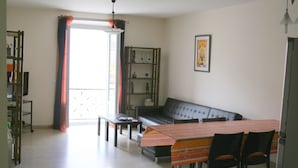 A living room with a sofa, a dining table with chairs, a bookshelf, and a window with curtains.