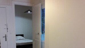 A hallway with a bed in the background, a trash bin, and a door leading to another room.