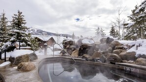 Une zone de sources chaudes où un panneau indique une profondeur de 3 pieds, entourée de rochers et d’arbres couverts de neige.