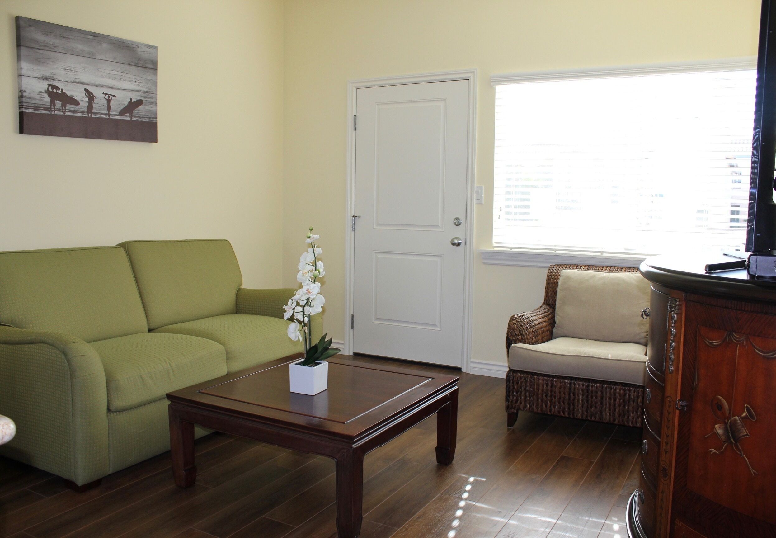 Una sala de estar con un sofá verde, una mesa de centro de madera con un jarrón blanco, una silla de mimbre y un aparador de madera.