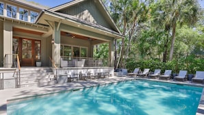 A swimming pool with lounge chairs, a covered patio, and a house with large windows.