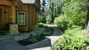 A wooden cabin with a stone fireplace, surrounded by a gravel driveway and lush greenery.