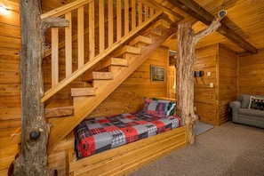 A log cabin interior with a wooden staircase, a bed with a red and gray quilt, and a gray sofa. - Perfect location, Secluded, Pet Friendly, Wooded Paradise (Sevierville)