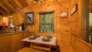 A wooden cabin interior with a dining table set for four, a kitchen area, and a large window with blinds. - Perfect location, Secluded, Pet Friendly, Wooded Paradise (Sevierville)