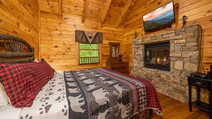 A log cabin bedroom with a stone fireplace, a bed with a red and black checkered blanket, and a mounted television. - Perfect location, Secluded, Pet Friendly, Wooded Paradise (Sevierville)