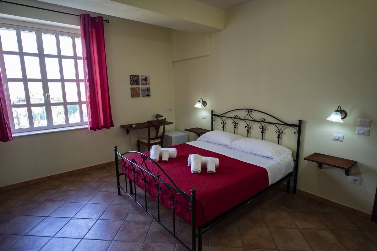 A bedroom with a bed, two bedside tables, a window with curtains, and a wall-mounted shelf.