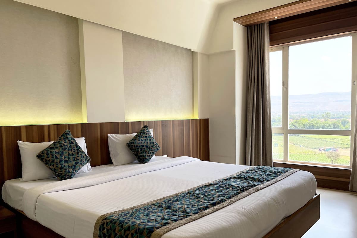 A hotel room with a large bed, two pillows, a headboard, and a window with a view of the countryside.