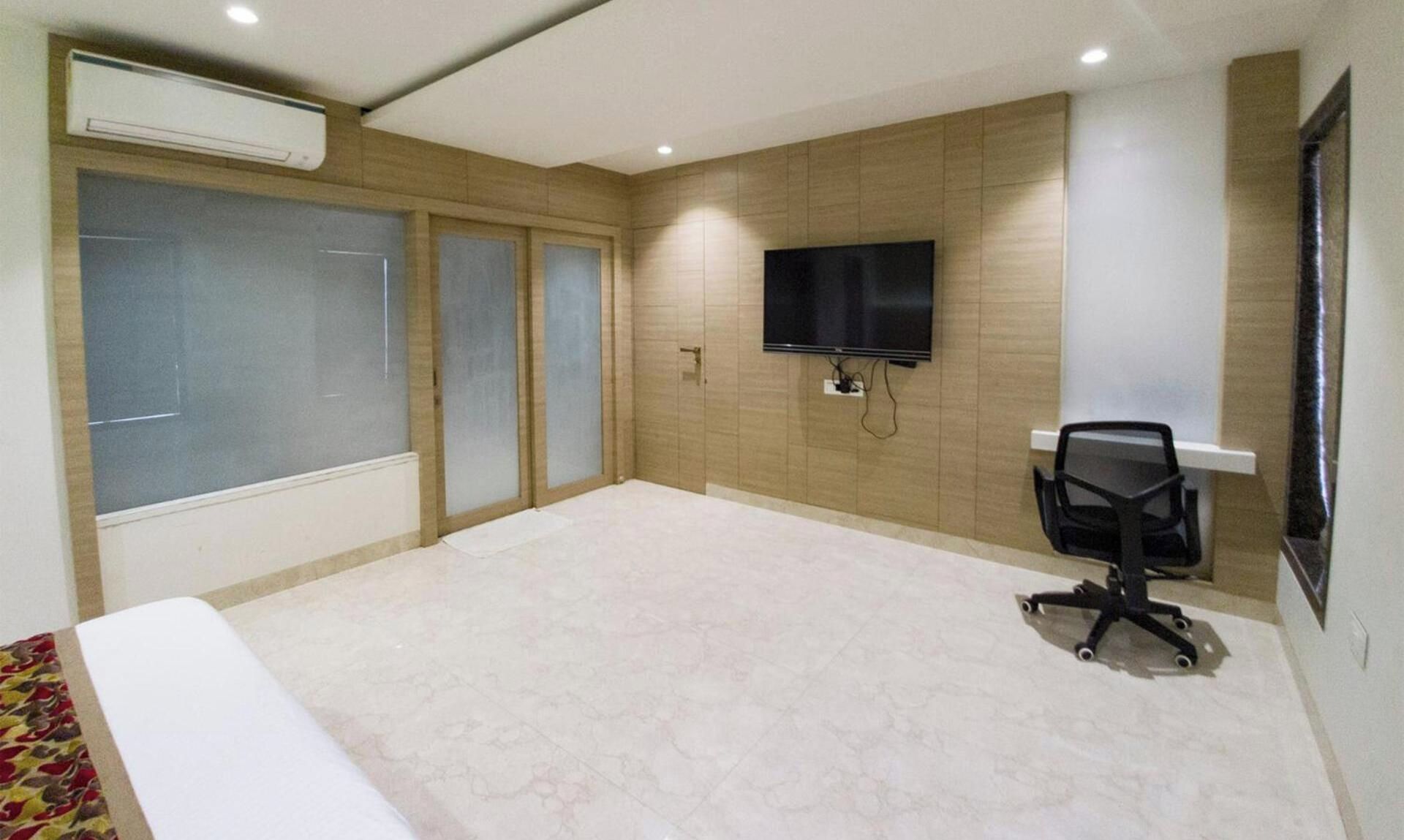 A hotel room with a large bed, a desk with a chair, a flat-screen TV mounted on the wall, and an air conditioning unit above the bed.