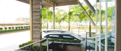 A covered outdoor area with a car parked inside, a staircase leading down, and a view of the street with parked cars and greenery.