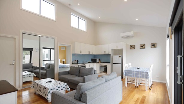 A spacious living area with a grey sofa, a dining table with chairs, and a kitchen area with white cabinets and appliances. - Apollo Bay Seal Apartments (Marengo)