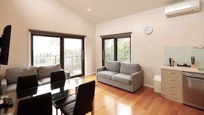 A modern living room with a dining table, two sofas, a kitchen area with a dishwasher, and large windows providing natural light. - Apollo Bay Seal Apartments (Marengo)