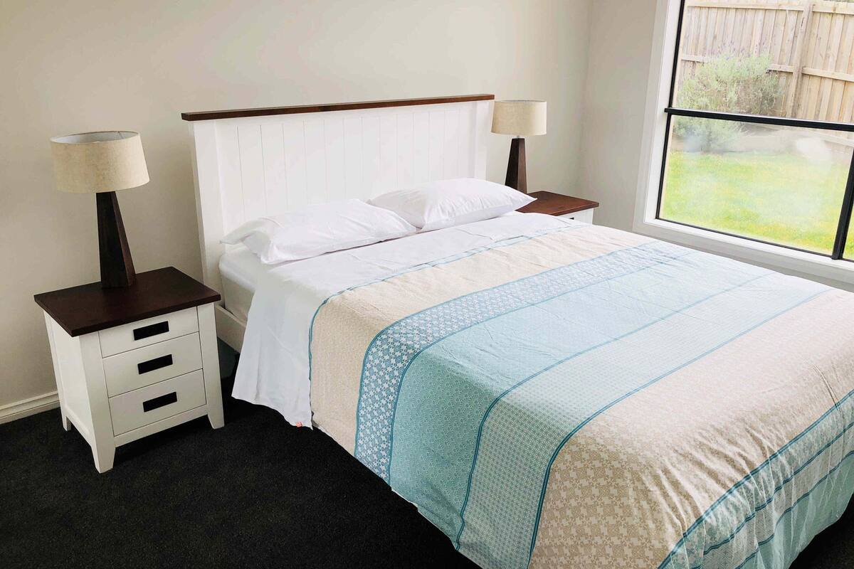 Una cama bien hecha con ropa de cama blanca y una manta estampada en azul y beige. Hay dos mesitas de noche con lámparas, y una ventana con vistas a una valla de madera y follaje.