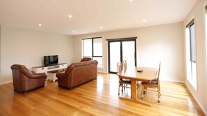 A spacious living room with a wooden floor, a dining table with chairs, a brown leather sofa, and a TV on a white cabinet. - Apollo Bay Seal Apartments (Marengo)