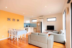 Una sala moderna con sofá, una mesa de comedor con sillas, una zona de cocina con armarios y electrodomésticos, y un televisor.