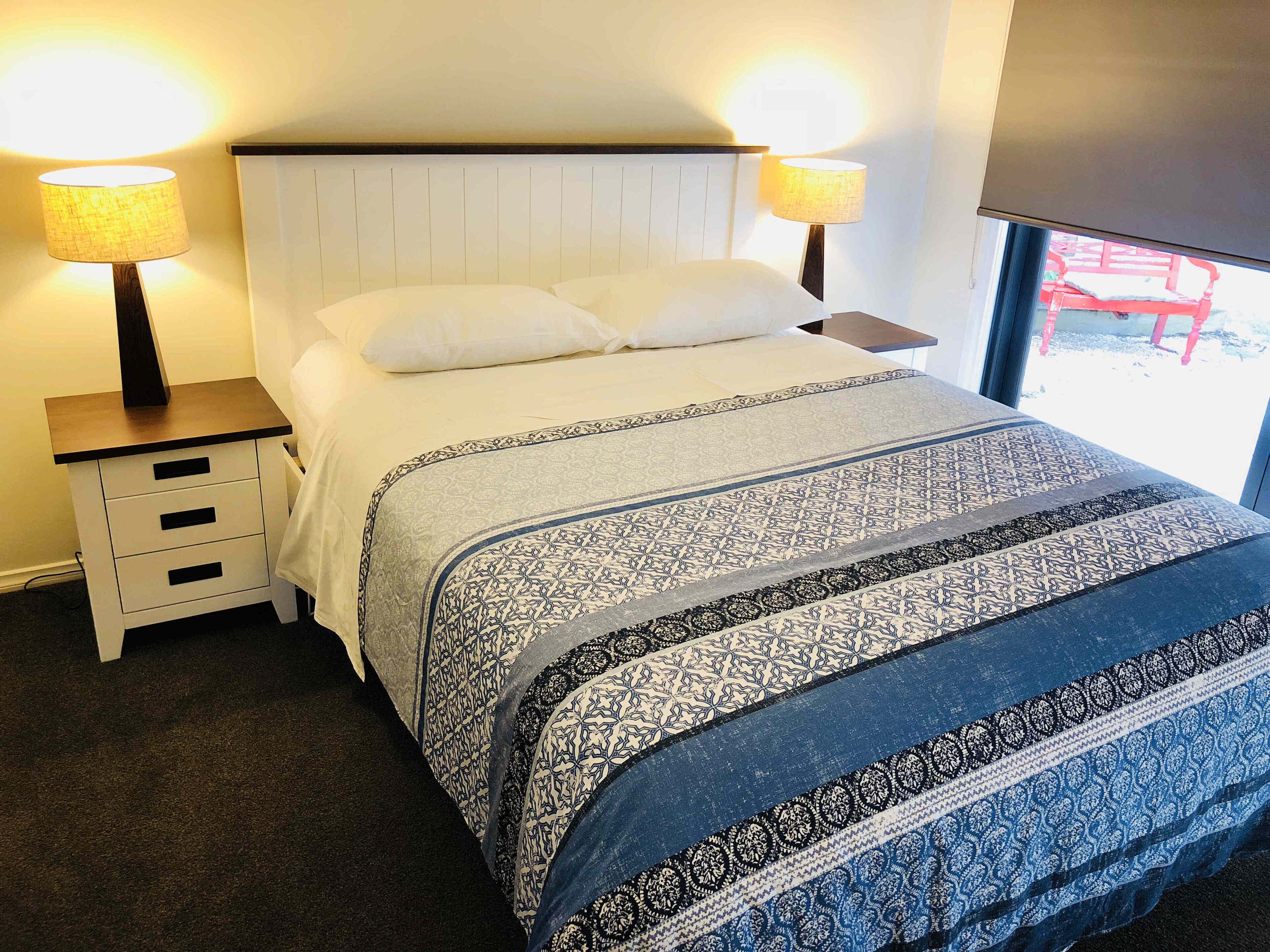 A neatly made bed with patterned bedding, two bedside lamps, a nightstand with drawers, and a window with a view of a red structure outside.
