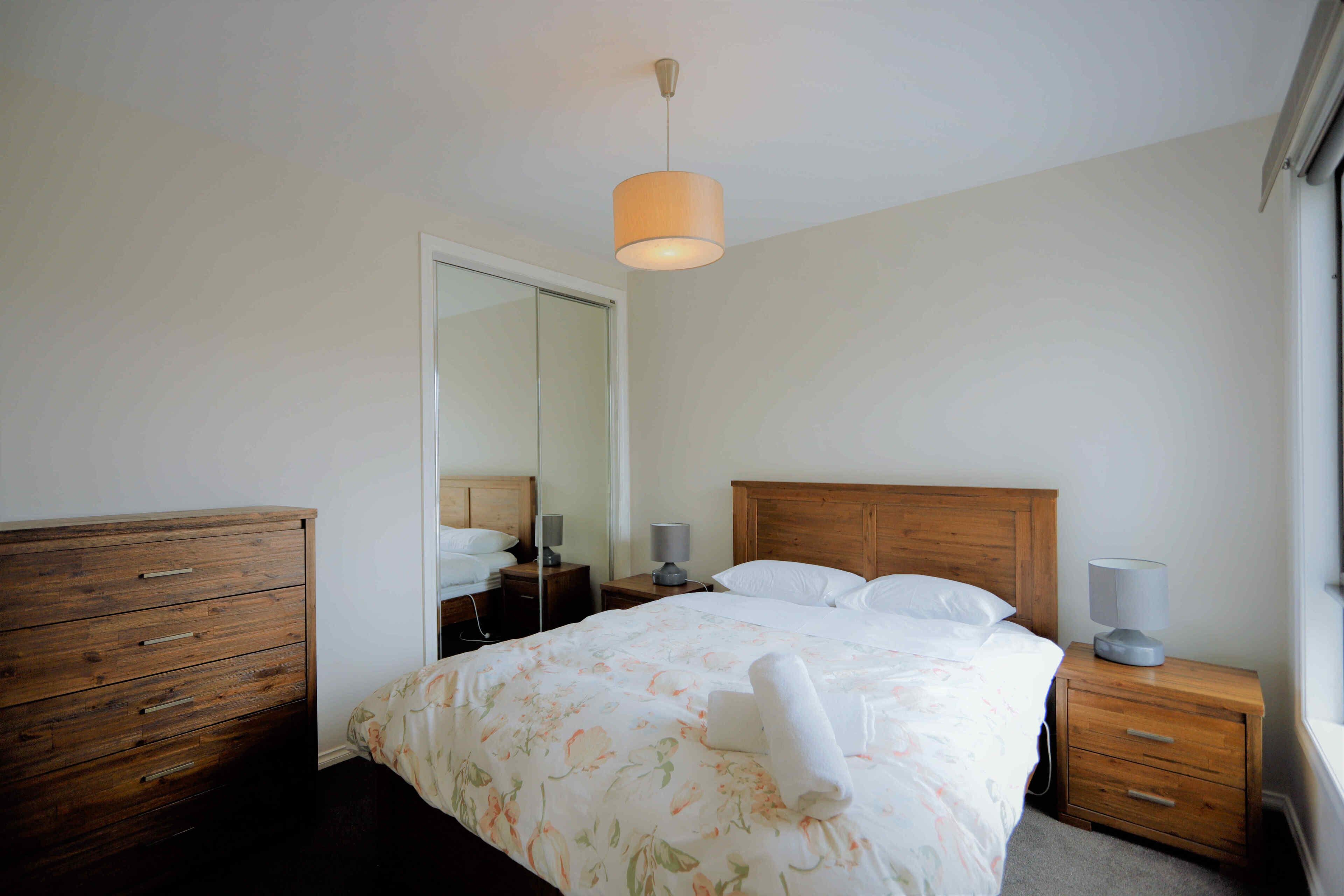 A bedroom with a wooden bed, a nightstand with a lamp, a dresser, and a mirror.
