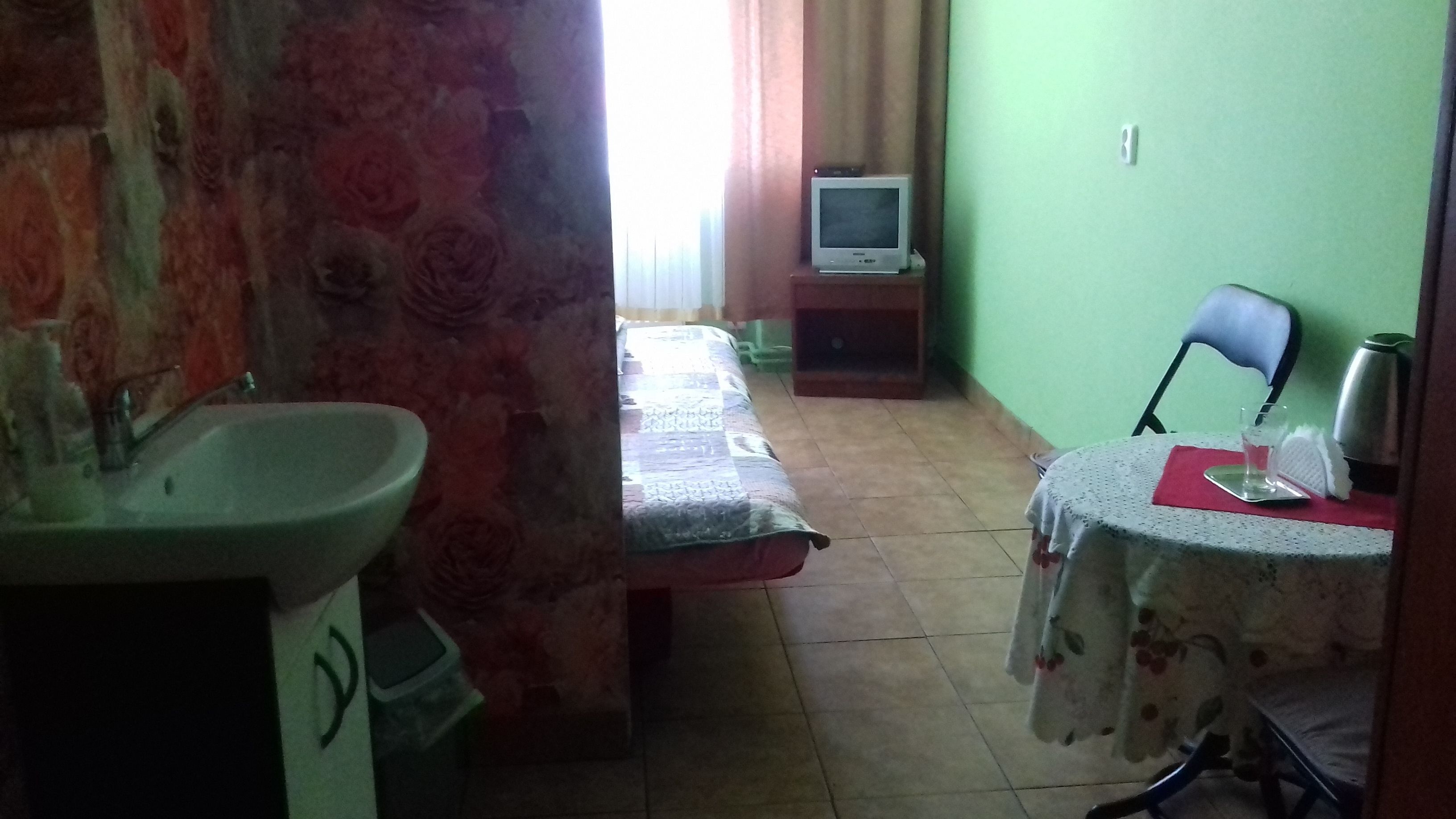 A small, simple room with a bed, a TV on a stand, a table with a kettle, and a sink.
