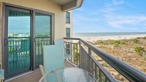 Ein Balkon mit Glastür, Tisch und Stühlen, mit Blick auf einen Strand mit Zaun und Bepflanzung.