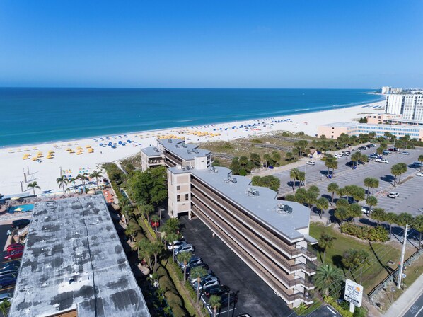 Une vue aérienne d’un hôtel avec un parking, d’une plage parsemée de parasols et d’un océan bleu azur.