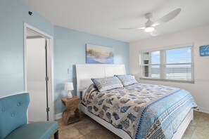 A bedroom with a bed, a chair, a ceiling fan, a window with blinds, and a painting on the wall.
