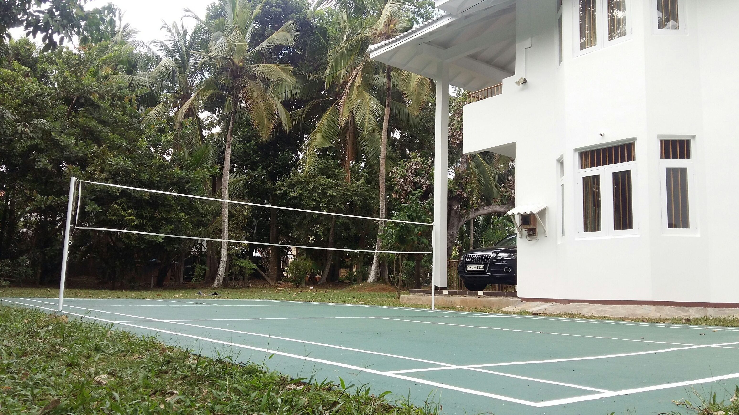 Un court de tennis avec un filet, entouré d’arbres et bordé d’un bâtiment blanc doté de fenêtres.