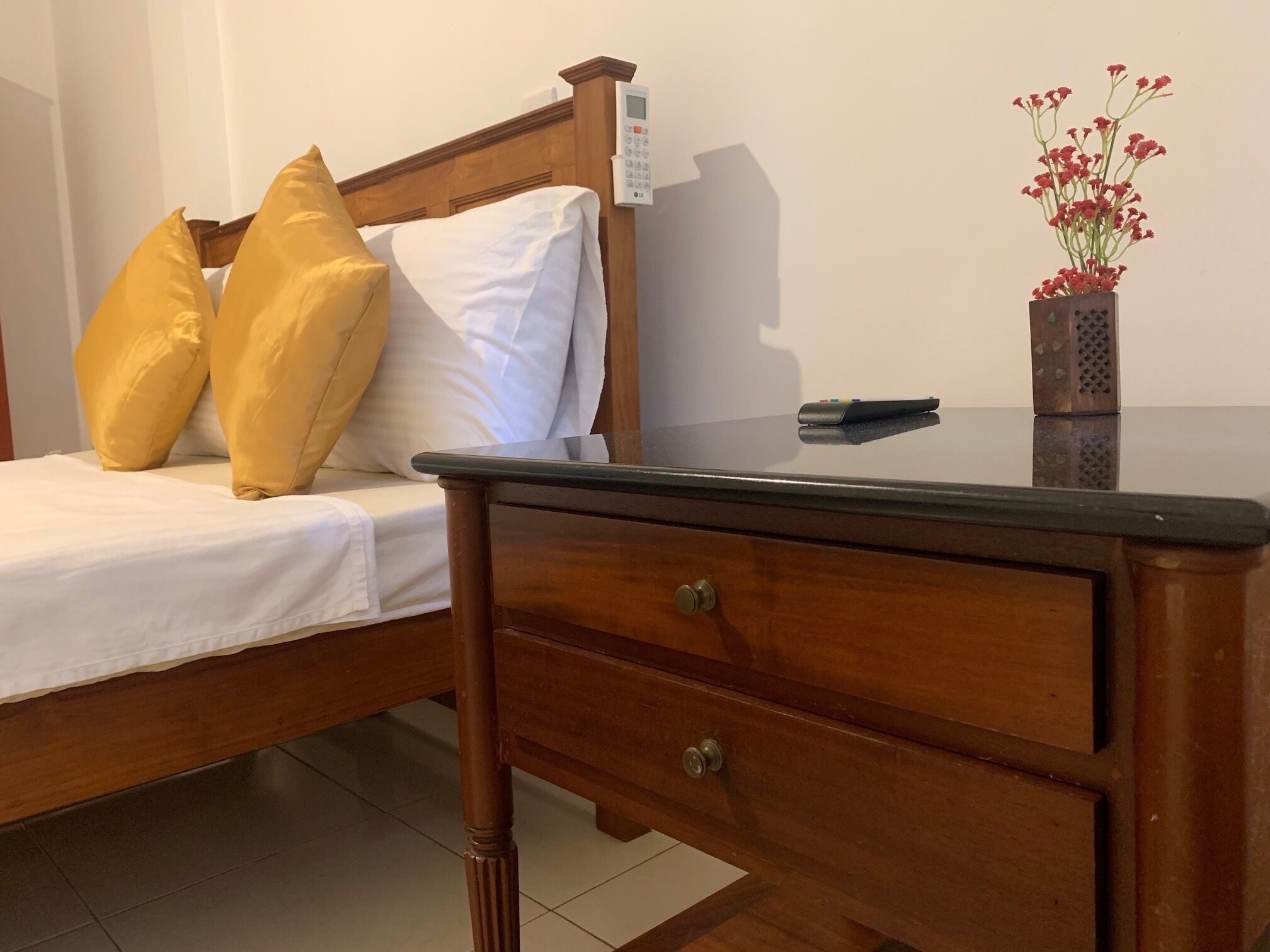 A wooden bed with white bedding, two yellow pillows, a bedside table with a remote control, and a vase with red flowers.