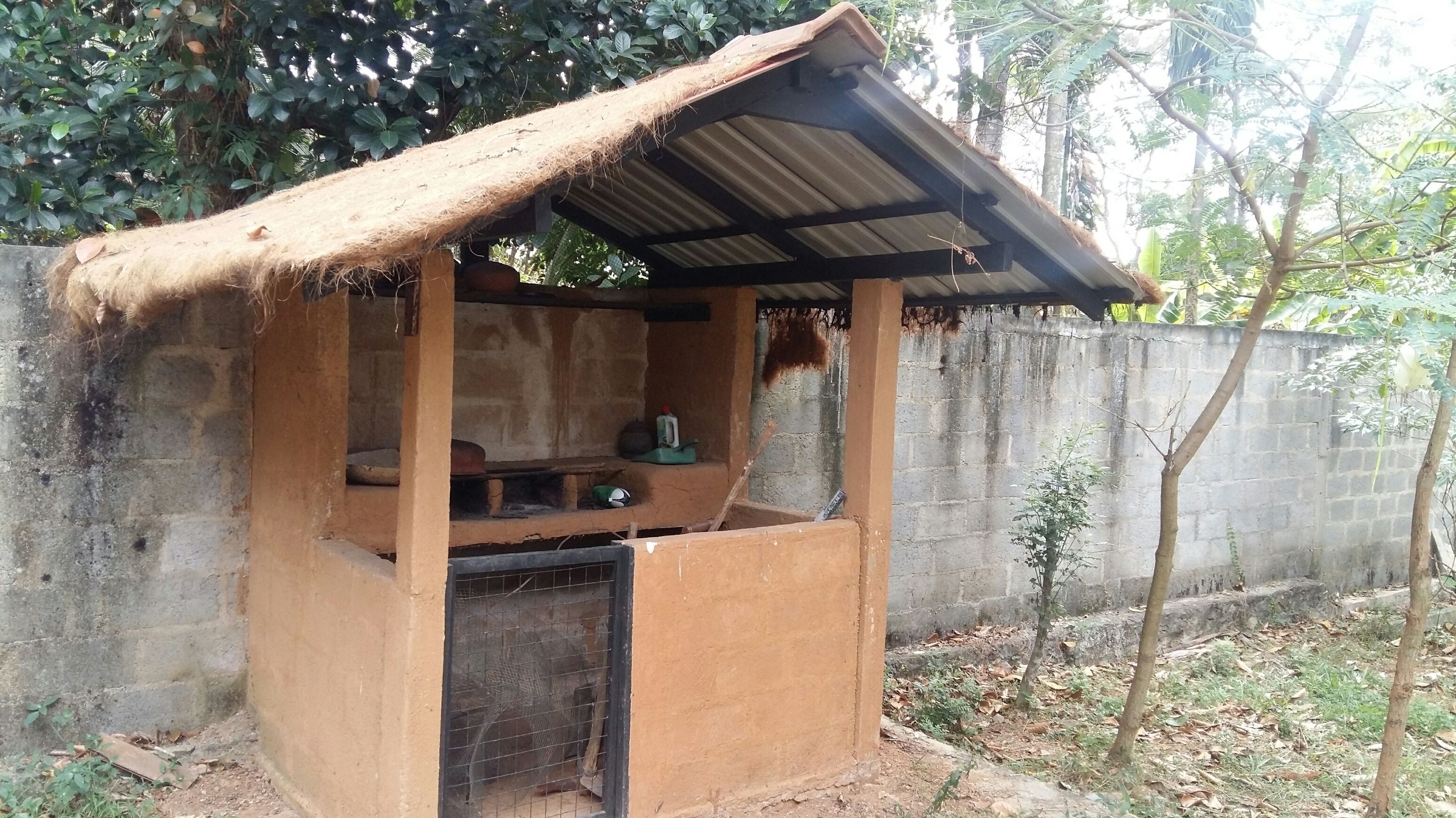 Una pequeña construcción rústica al aire libre con techo de paja y puerta de metal, rodeada de árboles y un muro de hormigón.