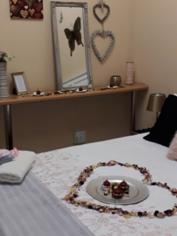 Habitación de hotel con cama, mesita de noche con espejo, una decoración con forma de corazón, y una pequeña mesa con un arreglo floral en forma de corazón y una vela.