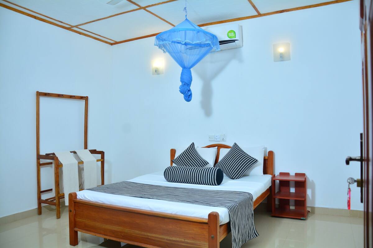 A bedroom with a wooden bed, a mosquito net, a bedside table, and a wall-mounted air conditioner.
